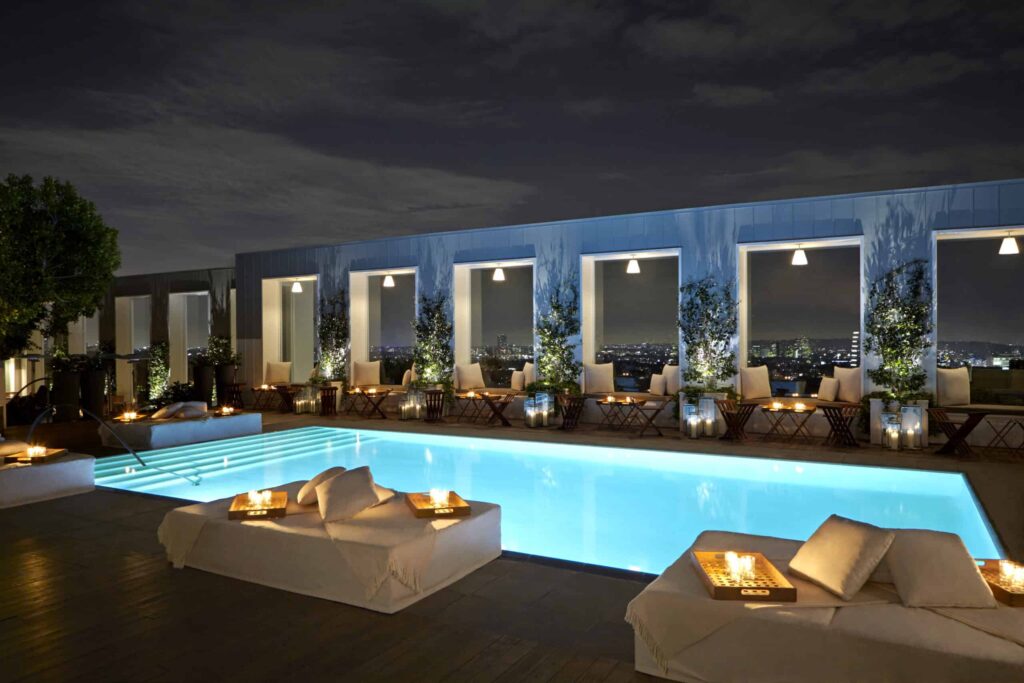 Outdoor rooftop pool at night at the Valorian Los Angeles, surrounded by lit candles, cushioned lounge chairs and city lights visible in the background. West Hollywood, CA