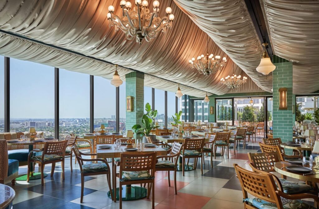Spacious restaurant with large windows, green tile pillars, chandeliers, wooden tables and chairs, and a view of the Los Angeles skyline. The Rooftop at The Sun Rose, West Hollywood, CA.
