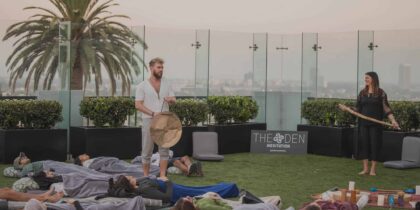 A meditation event by The Den Meditation on the rooftop of The London West Hollywood in CA, with two instructors and guests lying on cushioned mats surrounded by potted plants.