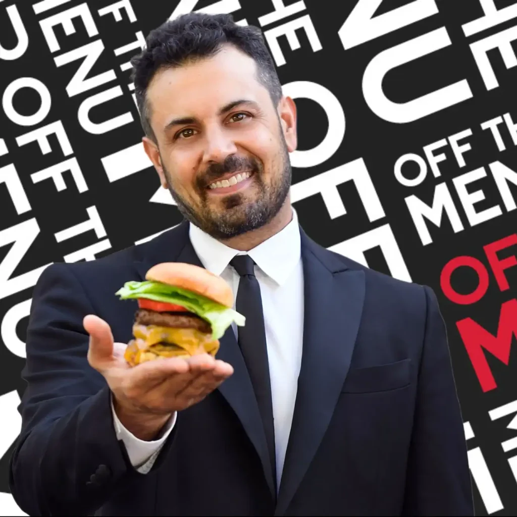A man in a black suit holds a cheeseburger with a beef patty, lettuce and tomato. The background features the repeated text "OFF THE MENU" in bold white and red letters.