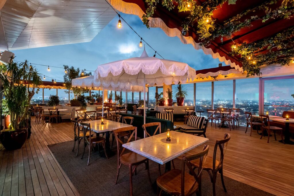Rooftop dining area at Harriet’s in West Hollywood, CA, with wooden floors, marble tables, string lights, plants and views of Los Angeles at dusk, with white umbrellas and foliage throughout the space.