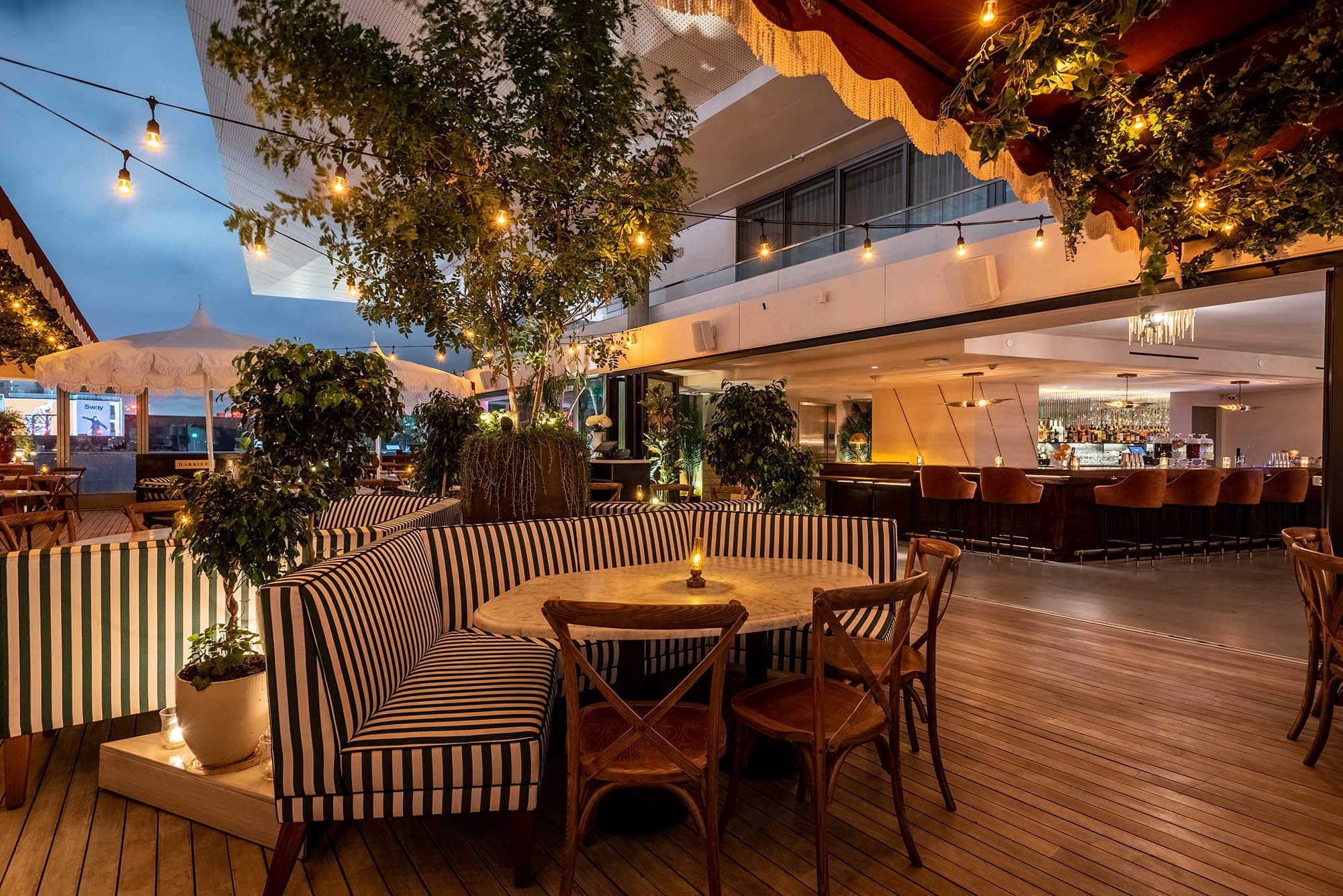 Outdoor restaurant seating area with striped benches, wooden tables and chairs, string lights, potted plants, and a bar area in the background.