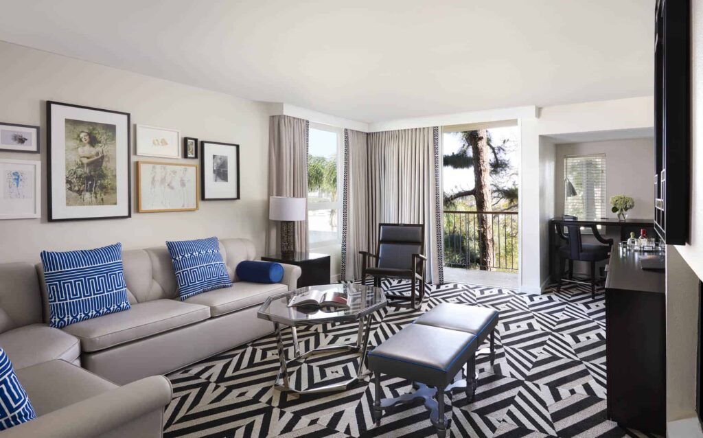 Modern living room at Chamberlain West Hollywood in CA with black-and-white patterned rug, beige sofa with blue pillows, glass coffee table, wall art, chairs and large windows letting in natural light.
