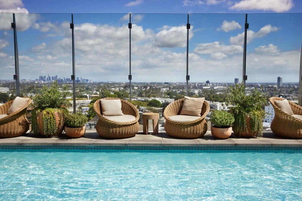 Wicker chairs and potted plants arranged in front of a glass-paneled wall on the rooftop at 1 Hotel West Hollywood in CA, with the Los Angeles skyline in the background.