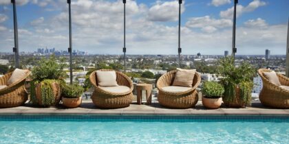 Wicker chairs and potted plants arranged in front of a glass-paneled wall on the rooftop at 1 Hotel West Hollywood in CA, with the Los Angeles skyline in the background.