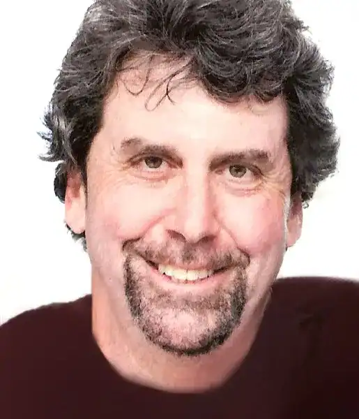 A middle-aged man with wavy dark hair, a short beard and a mustache is smiling at the camera against a plain background.