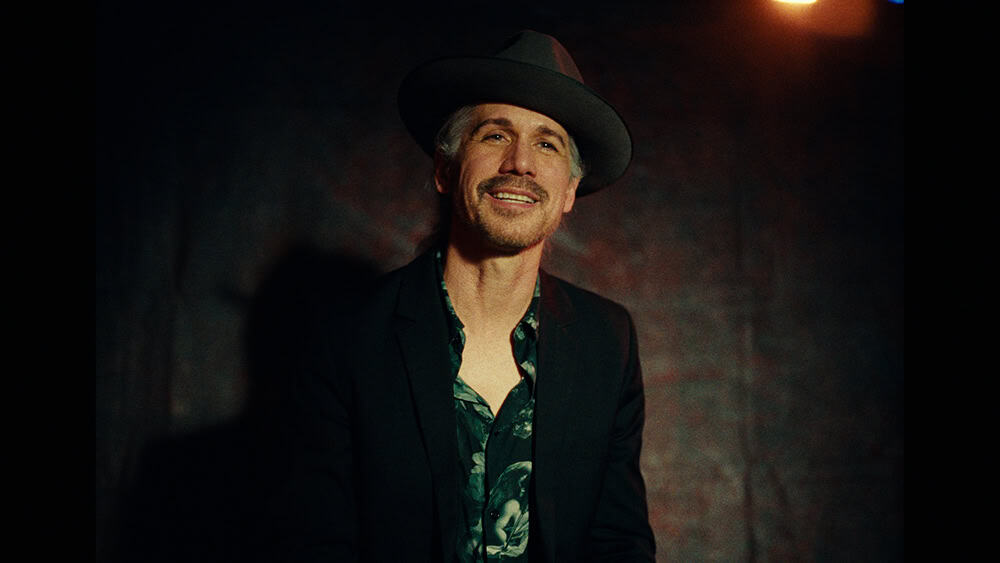 A man (Tommy Black) smiles while sitting in a room with moody lighting.