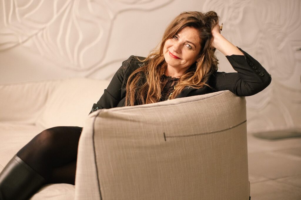 A woman dressed in black smiles at the camera while sitting on a white sofa with her head resting on her hand.