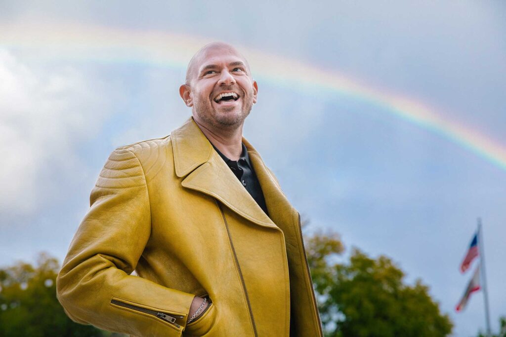 A man wearing a yellow leather jacket stands smiling with his hands in his pockets in front of a rainbow in West Hollywood, CA.