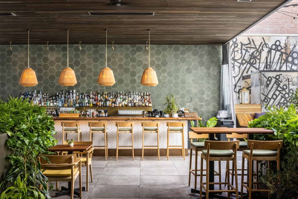 Modern bar interior at The Courtyard at Kimpton La Peer Hotel in West Hollywood, CA, with a hexagonal tiled wall, wicker pendant lights above a stocked bar, wooden chairs, tables and lush green plants.