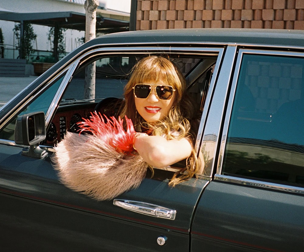 A woman (Mary Ta) leans out of a vintage car window wearing sunglasses.