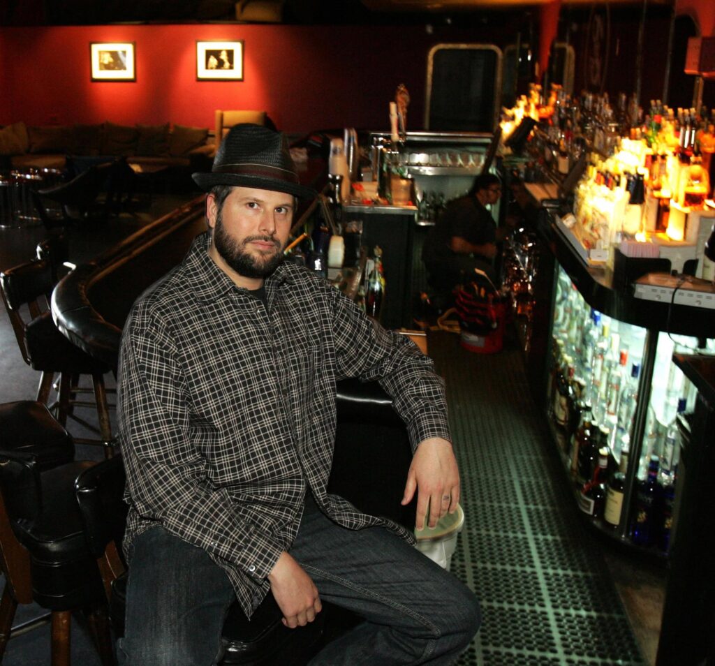 A man, Nic Adler, sits at a bar in West Hollywood, CA, wearing a black hat and plaid long-sleeve shirt.