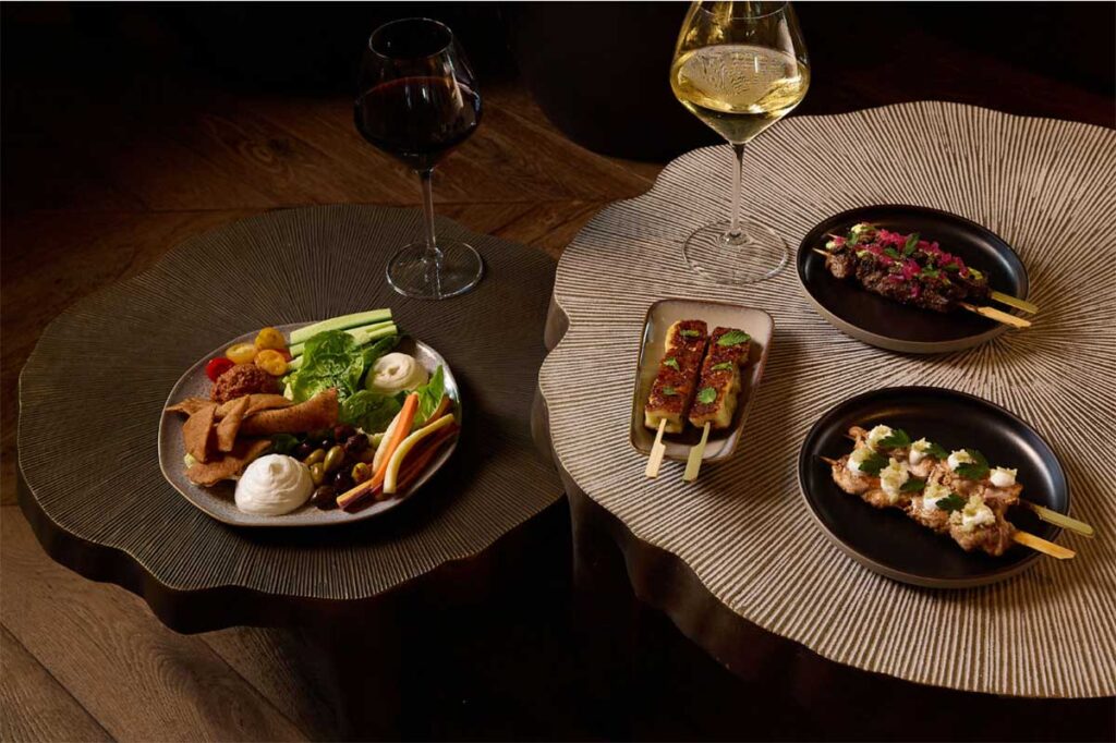 A snack platter with vegetables and dip, skewered appetizers and glasses of red and white wine arranged on textured round tables in a moody setting.