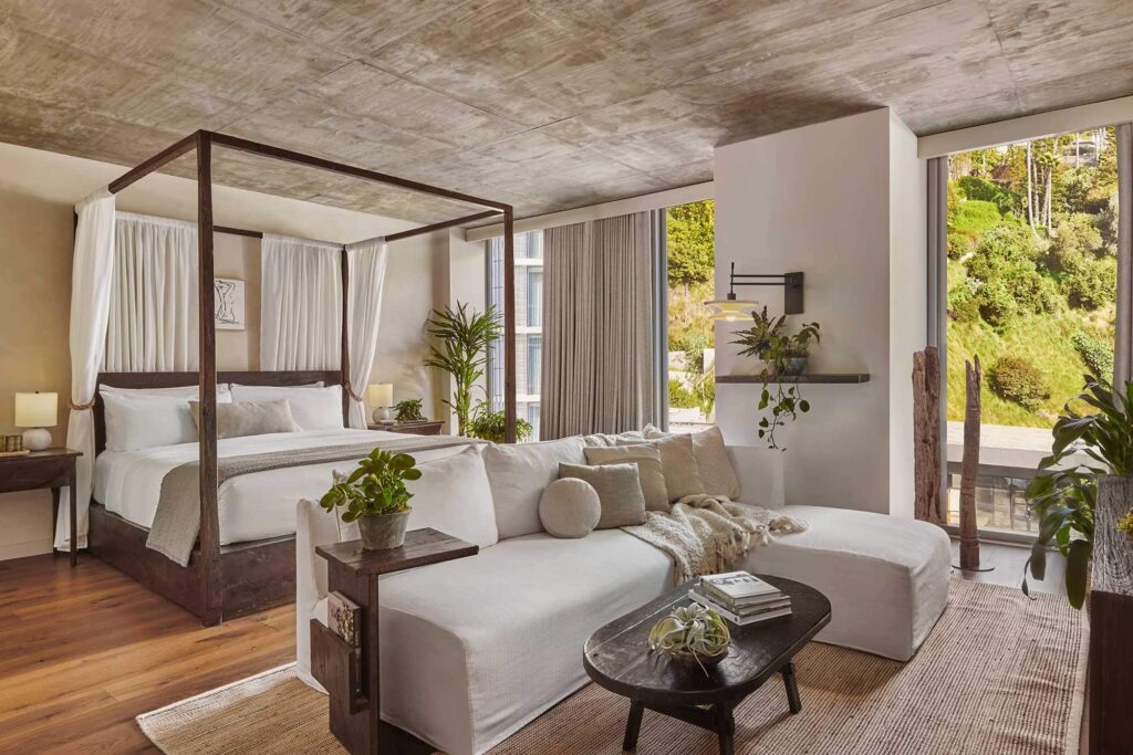 Interior of a Studio Suite at 1 Hotel West Hollywood in CA with a wooden canopy bed with white drapes, a neutral-toned sectional, wooden coffee table and beige curtains beside a window overlooking trees.