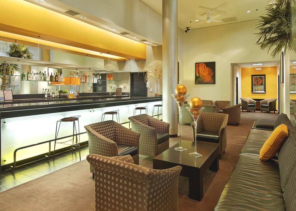Lobby area of Ramada Plaza in West Hollywood, CA, with yellow walls, patterned armchairs, a black leather sofa, a glass coffee table and a backlit bar with barstools, framed artwork and tall plants.