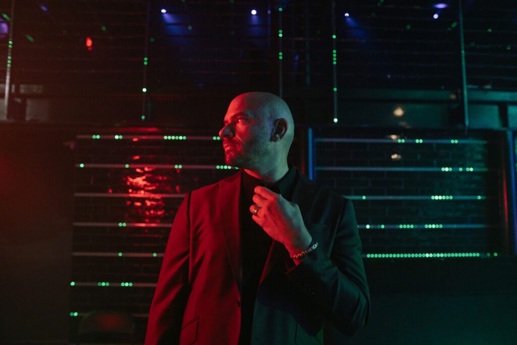 A man in a dark suit stands in a dimly lit room with colorful red and green lights, looking to the side.
