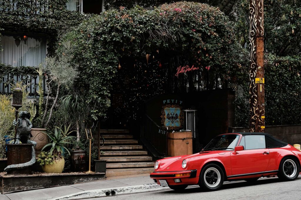 A red vintage car parked outside the foliage-covered entrance of Petit Ermitage