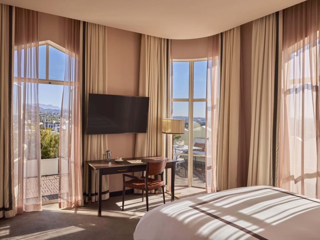 Hotel room at Sunset Tower Hotel in West Hollywood, CA, with large windows, a desk and chair, mounted TV and a white bed overlooking Los Angeles.