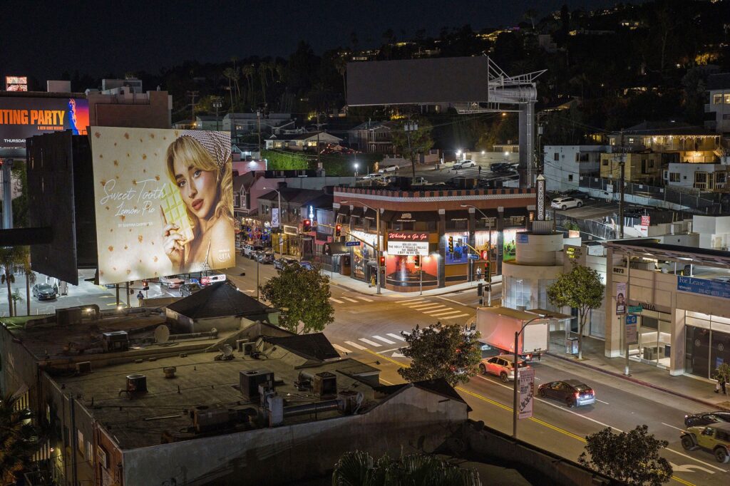 Aerail view of Sunset Strip in West Hollywood, CA, at night with illuminated streets, buildings and a large billboard featuring Sabrina Carpenter and the text "Sweet Tooth" above a busy intersection.