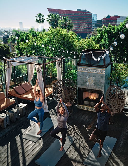 Three people do yoga on a rooftop in West Hollywood with Pacific Design Center visible in the background.