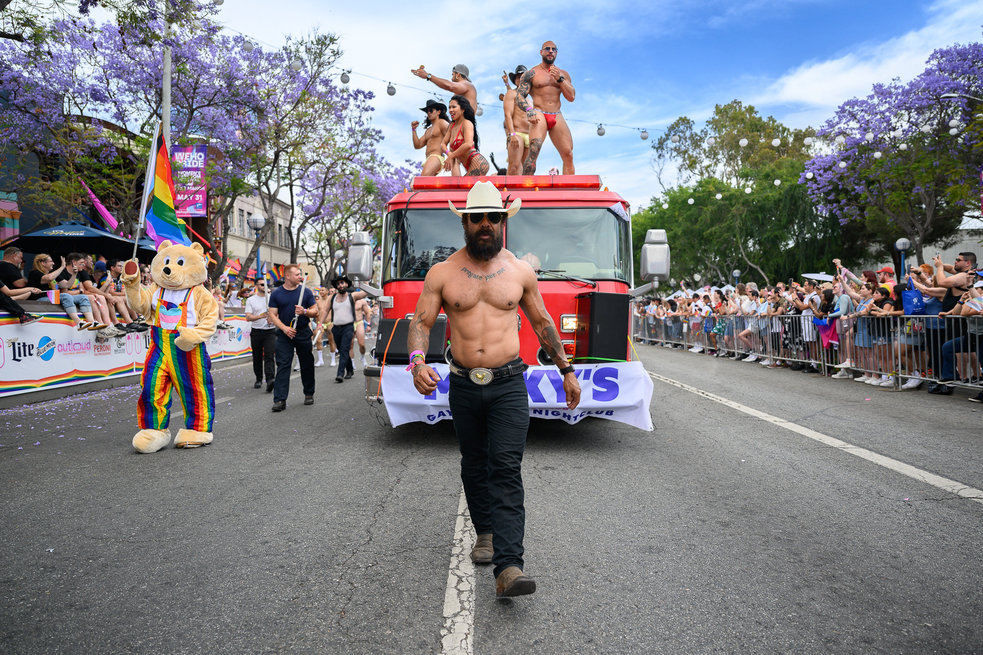 A firetruck carrying male go-go dancers, along with a shirtless man walking ahead, participates in the WeHo Pride festival.