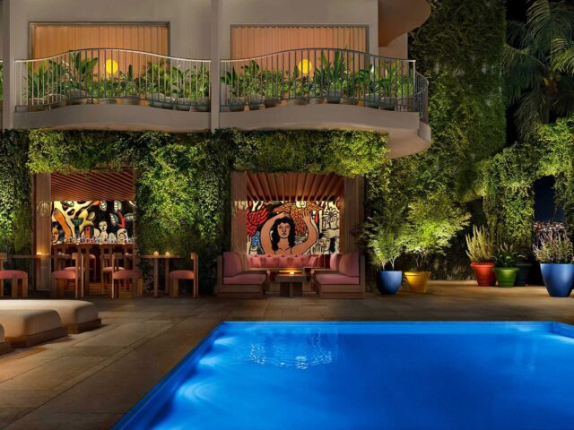 A poolside seating area at PUBLIC West Hollywood hotel at night with colorful murals, potted plants, and greenery covering the building's exterior; balconies overlook the illuminated scene.