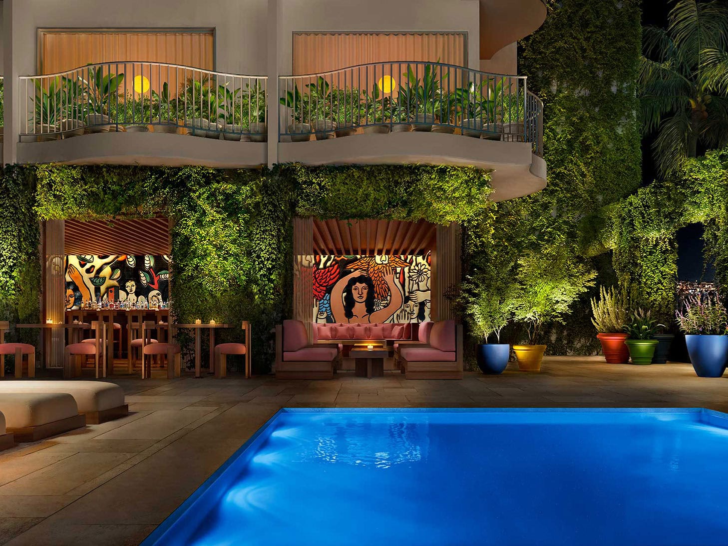 A poolside seating area at PUBLIC West Hollywood hotel at night with colorful murals, potted plants, and greenery covering the building's exterior; balconies overlook the illuminated scene.