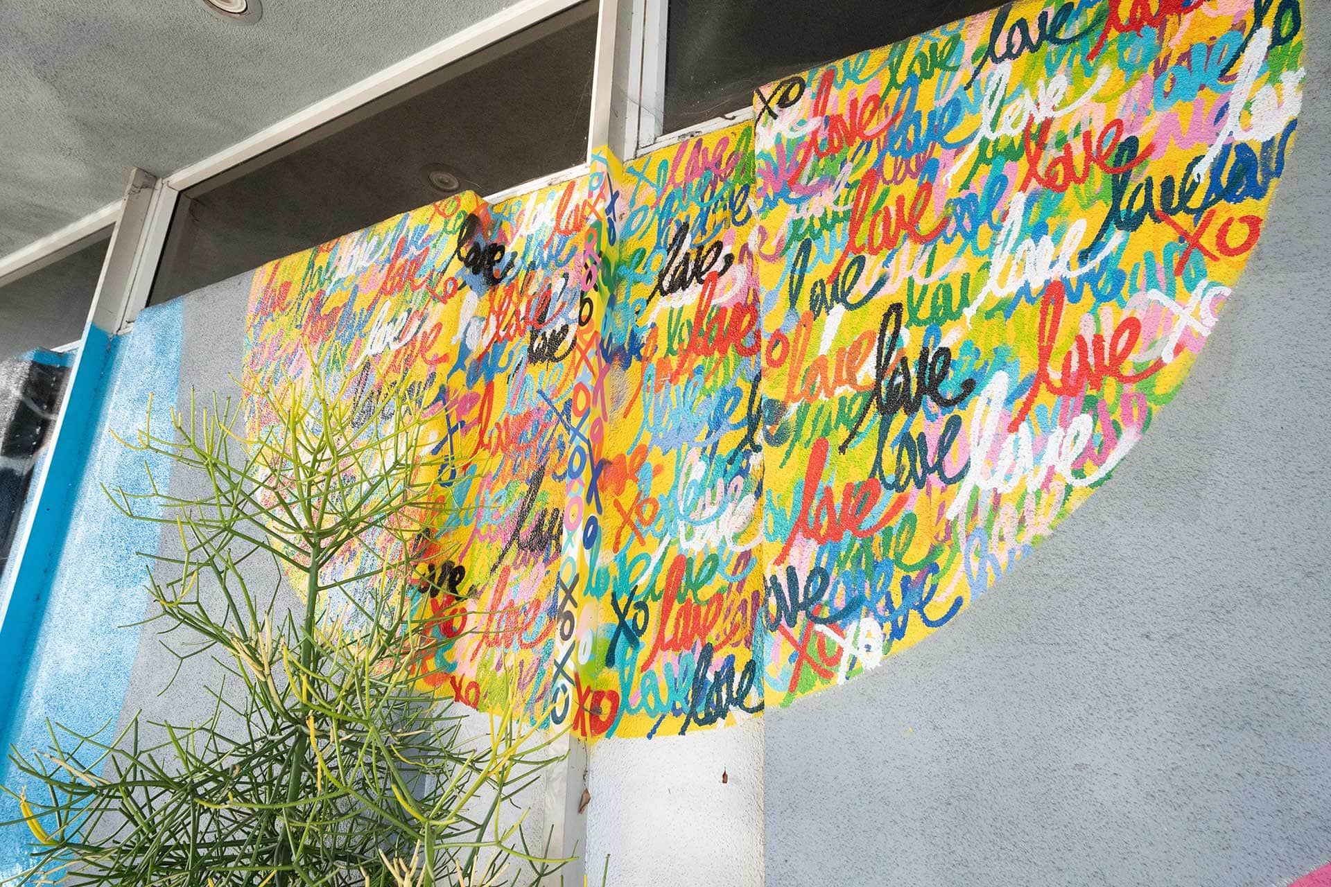 A mural on a wall features the word "love" repeatedly painted in colorful script, forming a shape resembling a tree. West Hollywood, CA