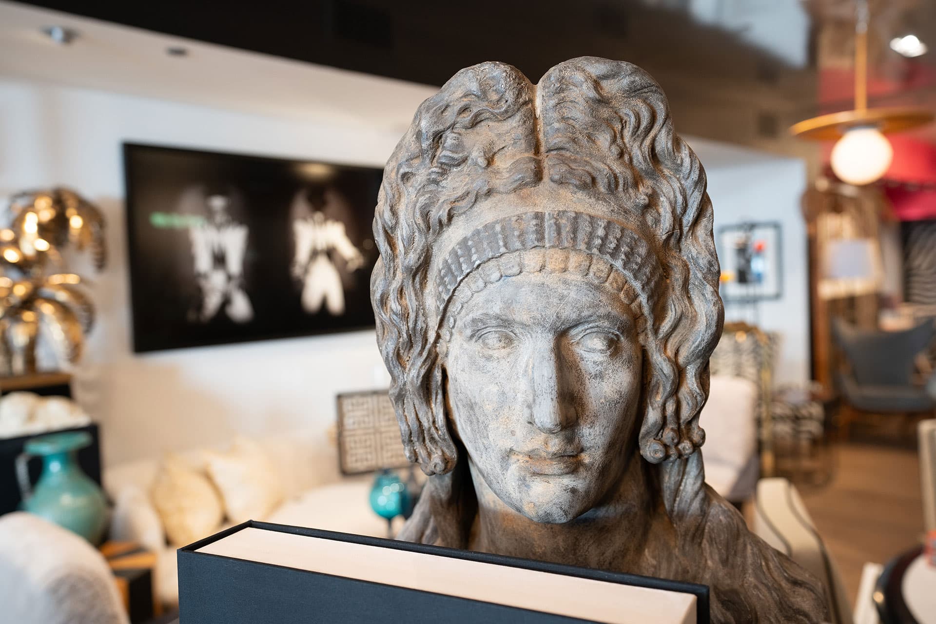 Stone bust of a figure with wavy hair and a headband, holding a book, displayed in a modern, well-lit room with eclectic decor.