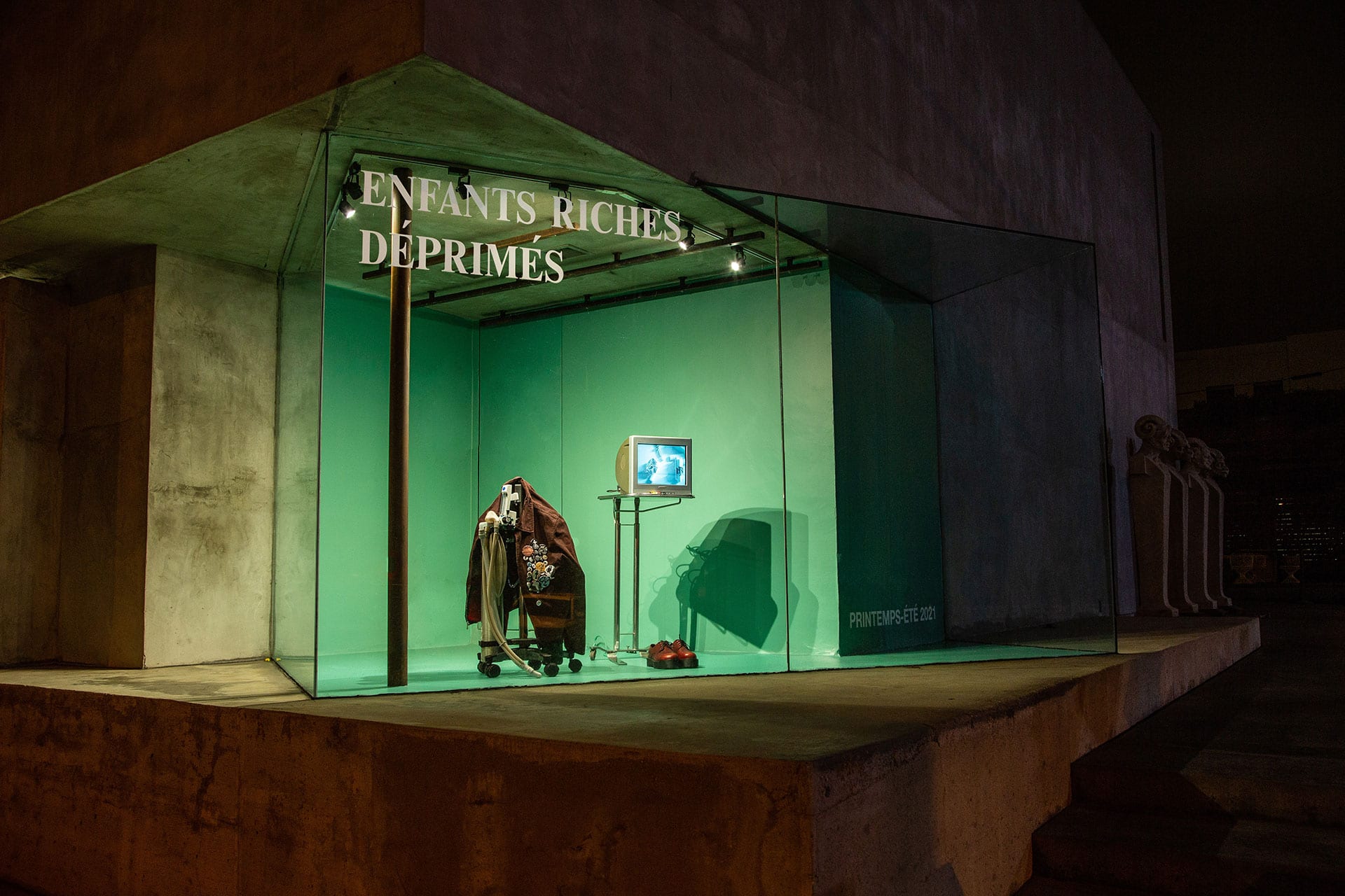 A glass display case with a green backdrop features hanging clothes, a TV on a stand, and the words “ENFANTS RICHES DEPRIMES” on the window at Maxfield in West Hollywood