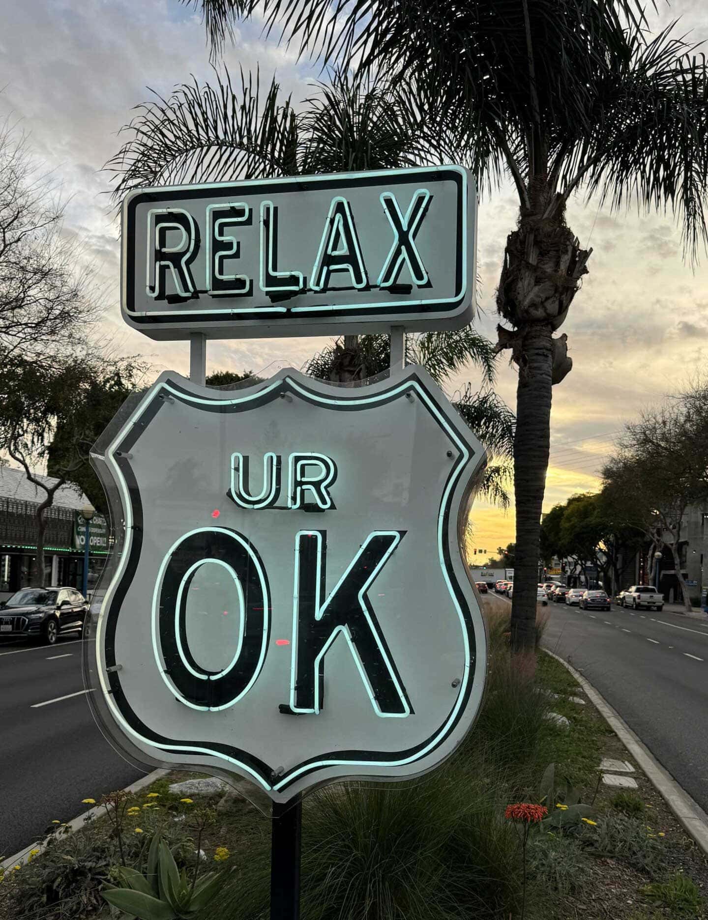 A neon sign on Santa Monica Boulevard in West Hollywood reads “RELAX” above a shield-shaped sign that says “UR OK,” with palm trees and cars on a street in the background at sunset.