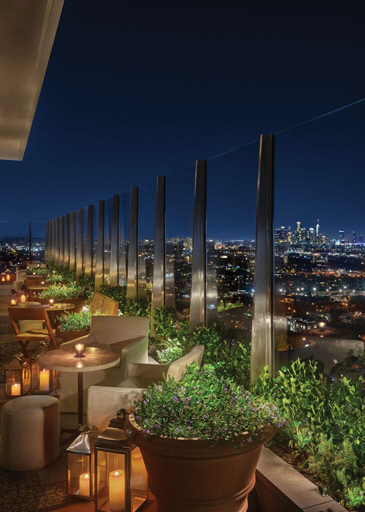 Outdoor rooftop lounge at night with city skyline view, potted plants, candlelit tables, cushioned chairs, and glass railing.